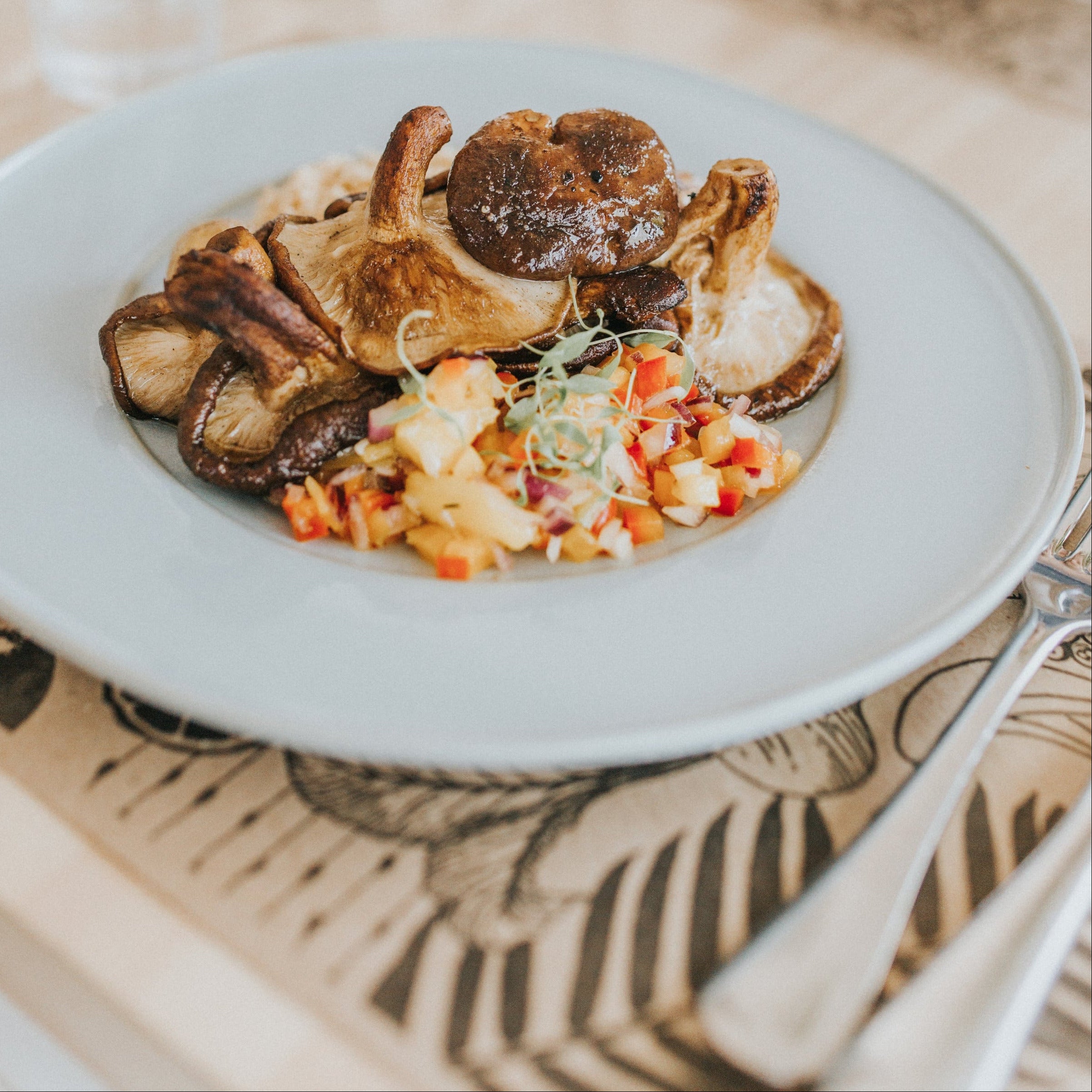 A plated dish of roasted mushrooms and a colorful vegetable rice salad on a light gray plate, set on a patterned table runner at a dining table.